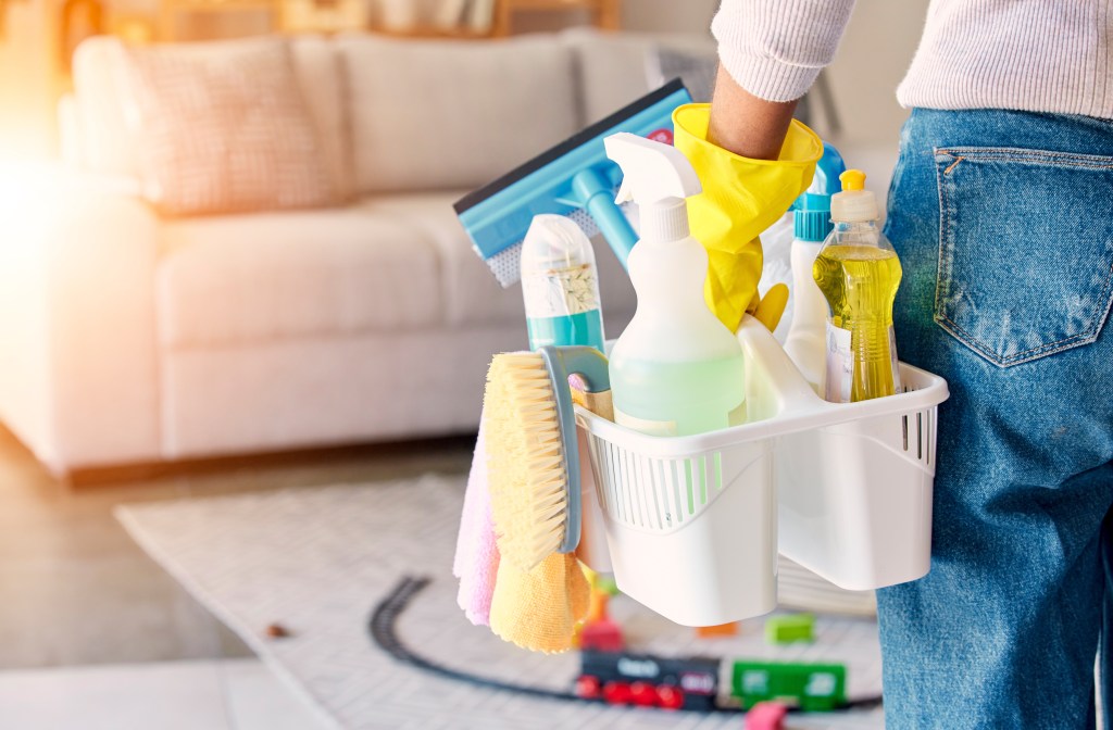 A person wearing yellow gloves and jeans carries a basket of cleaning supplies, including brushes, bottles, and cloths, with a blurred living room in the background.