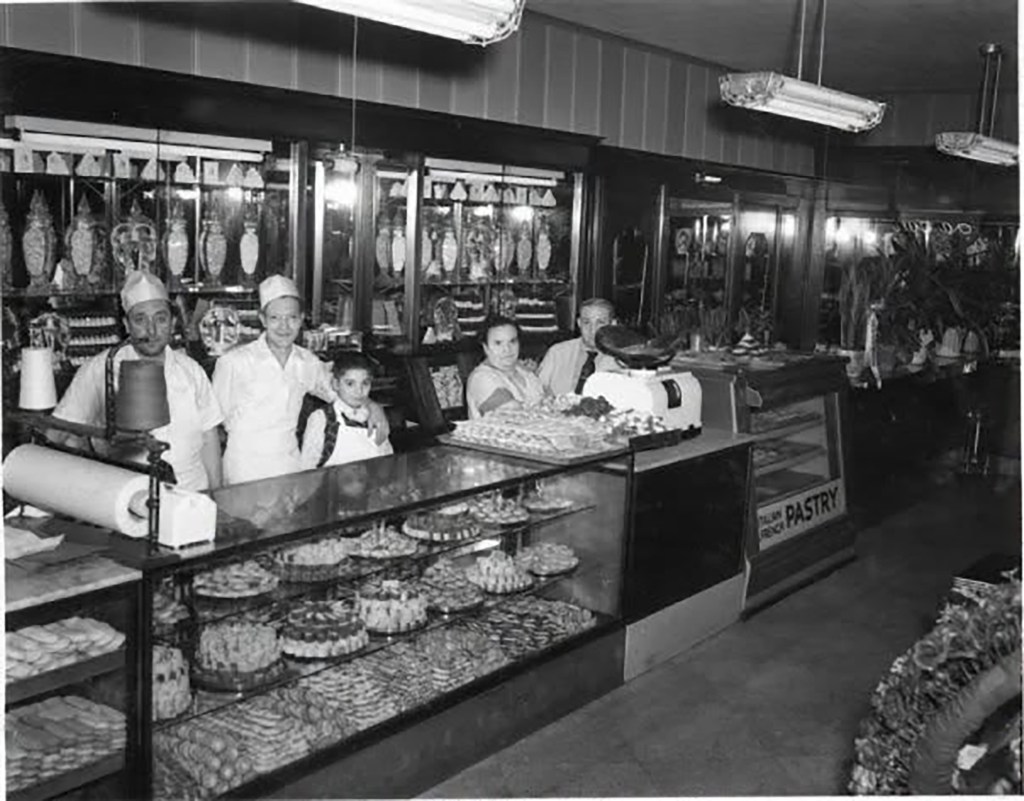 Black and white photo of four adults and one child behind a bakery counter filled with pastries.