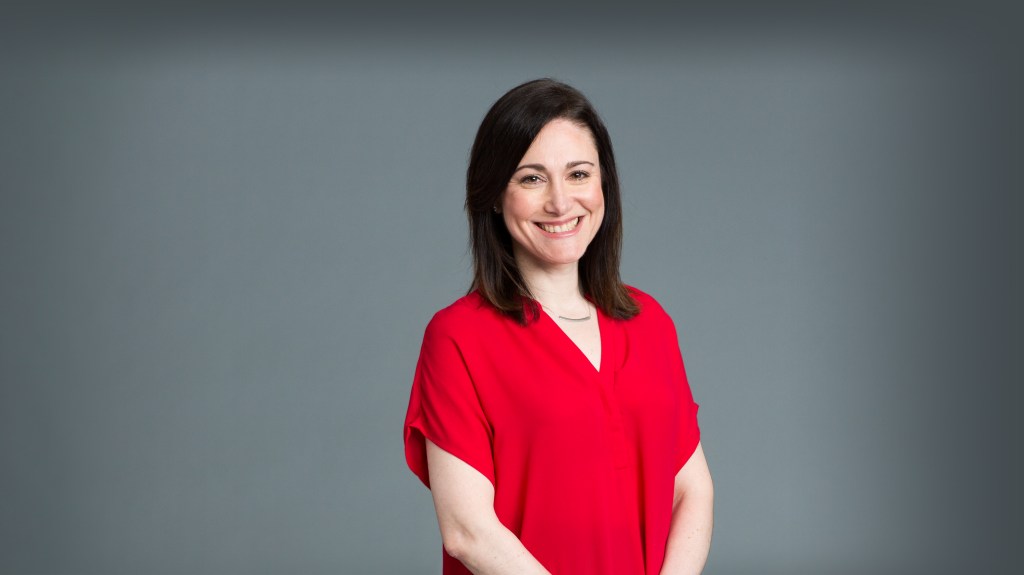 Melanie K. Greifer, a woman with dark hair and a red top, smiles at the camera.