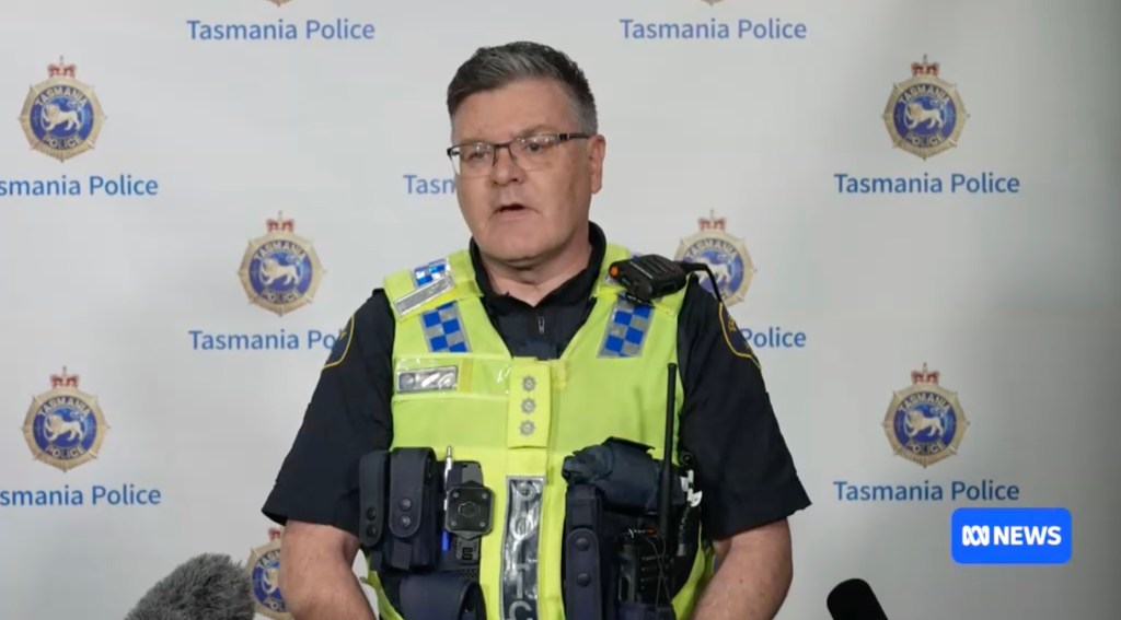 A police officer in a high-visibility vest speaks at a press conference with the "Tasmania Police" logo in the background.