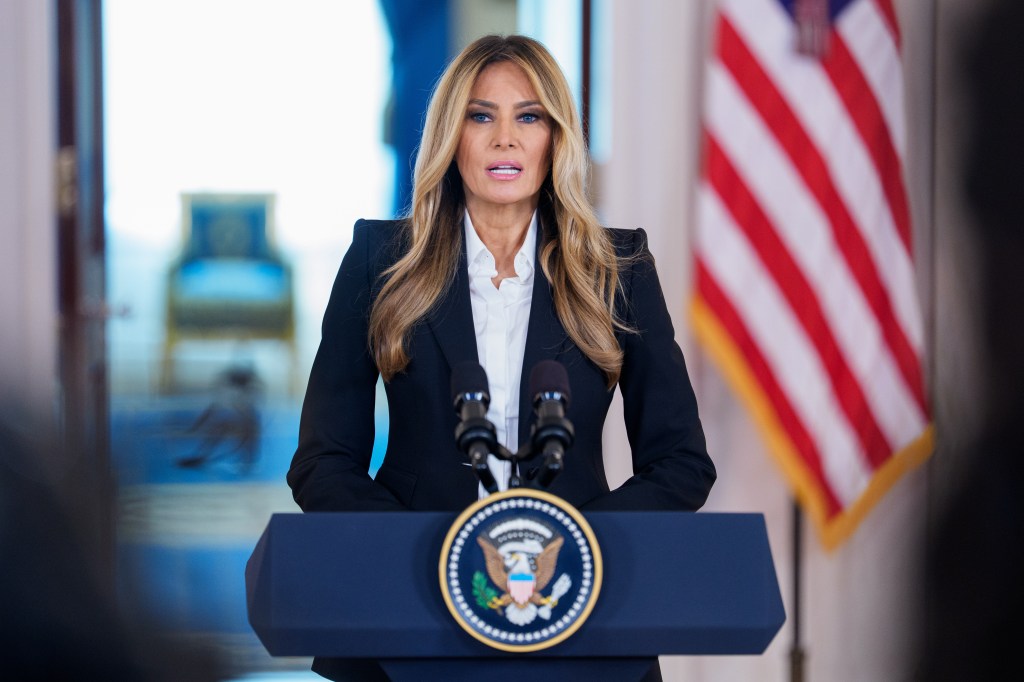 Melania Trump speaking at a podium with two microphones, an American flag in the background.