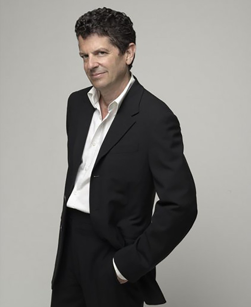 Michael Gross, a man with curly hair, in a black suit with an open white shirt, looks at the camera.