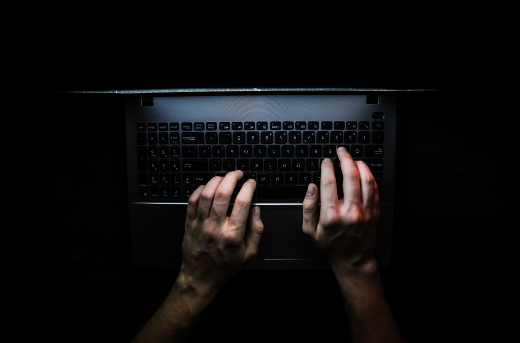 An overhead shot of a person's hands typing on a laptop keyboard in a dark room.