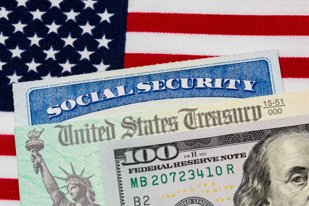 A Social Security card, United States Treasury check, and a $100 bill lie on top of an American flag, symbolizing social security benefits.