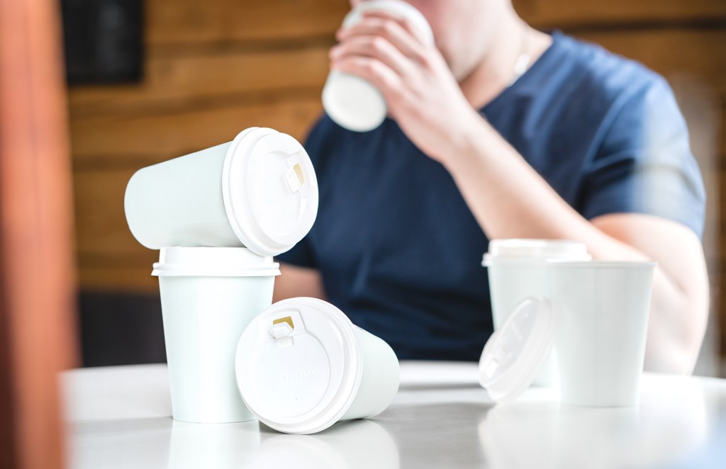 A man in a dark blue shirt drinks from a white coffee cup while several empty coffee cups with white lids are on the white table in front of him.