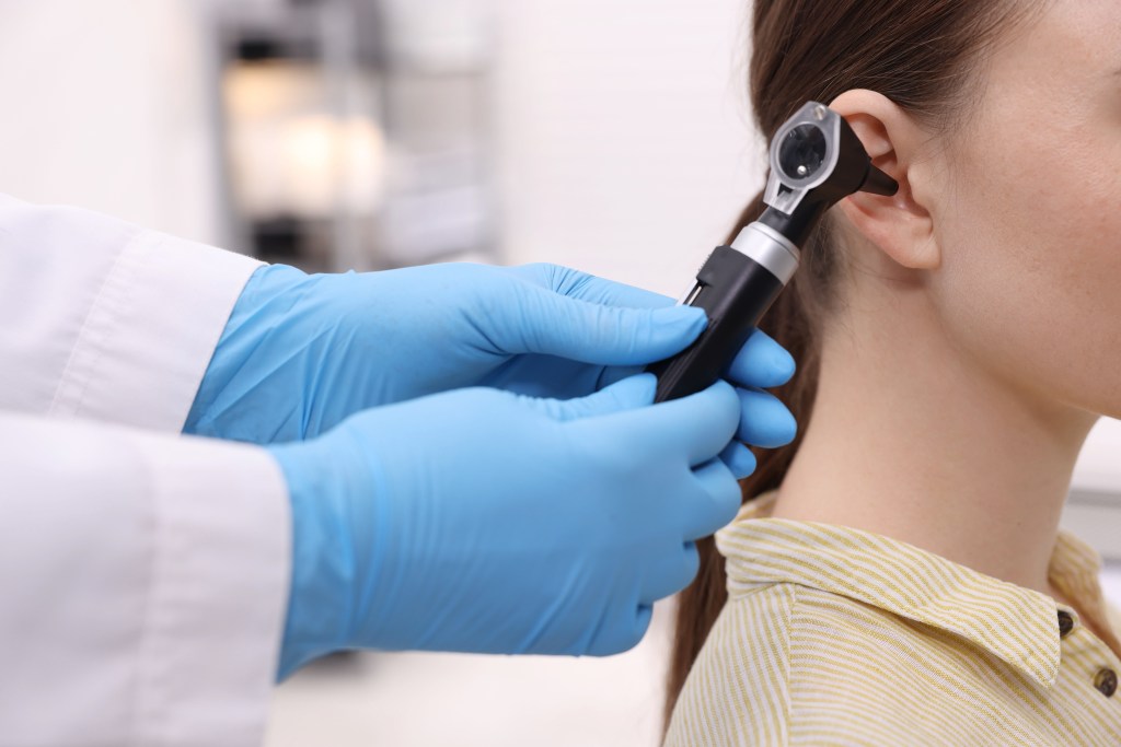 Doctor in blue gloves examines a patient's ear with an otoscope.