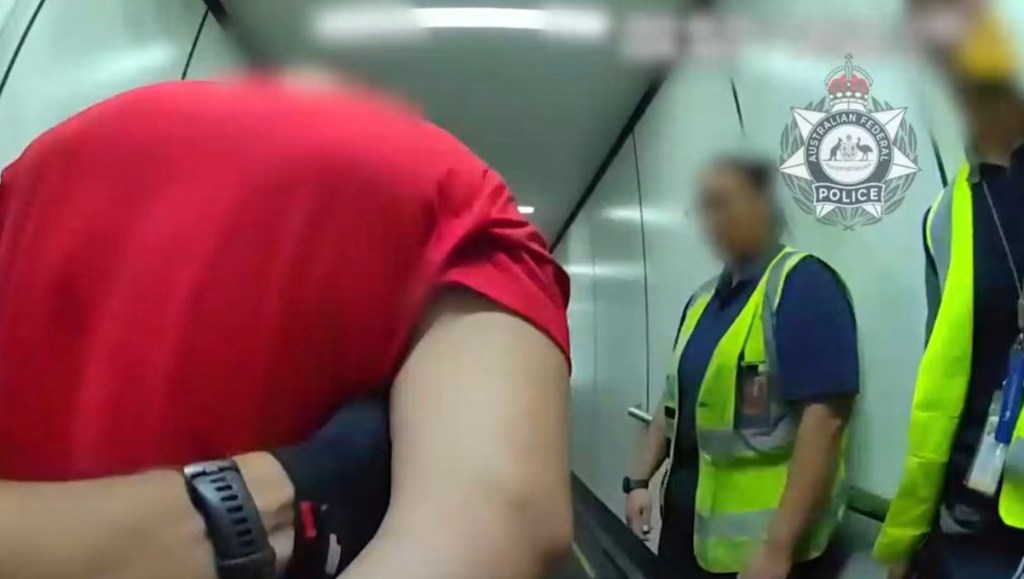 A blurry image of a man in a red shirt on an escalator with two uniformed Australian Federal Police officers.