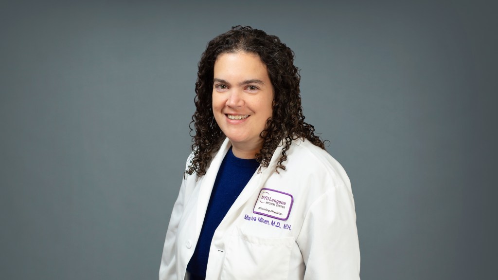 Dr. Mia Minen, a woman with dark curly hair and a white lab coat, smiles at the camera.