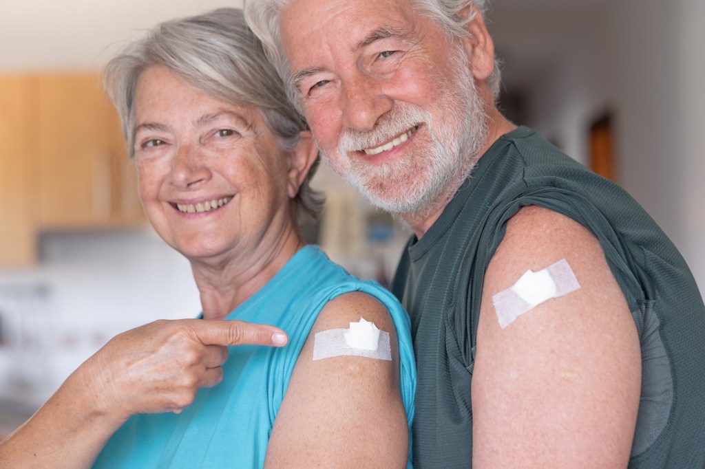 A smiling senior couple, a woman and a man, showing off their arms with bandages from their COVID-19 booster shots.