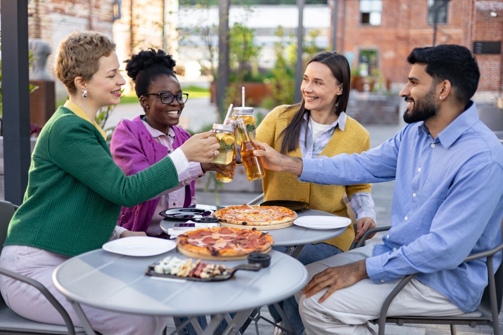 A diverse group of four friends toasting with drinks over pizzas at an outdoor table