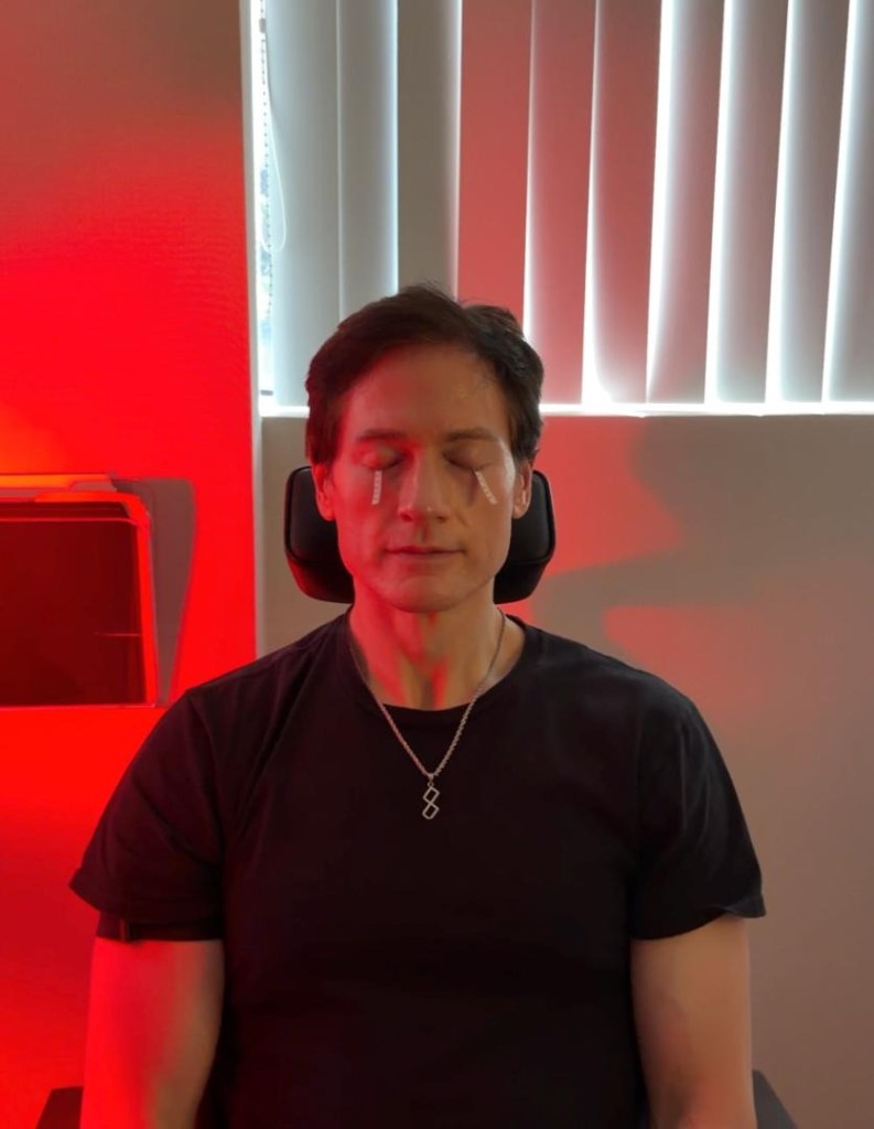 Bryan Johnson with patches under his closed eyes, sitting in red light therapy.