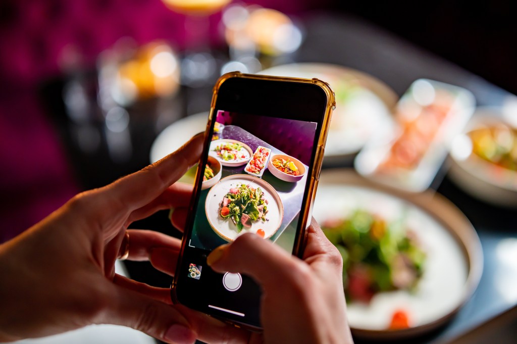 Woman holding a smartphone to photograph several plates of food at a restaurant.