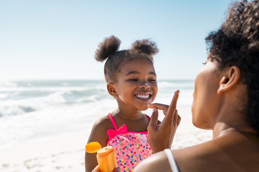 A black mother applying sunscreen to her daughter's nose at the beach.