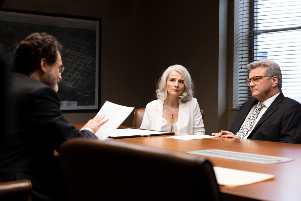 Three people in business attire sit at a conference table, with two looking toward the person on the left who holds papers.
