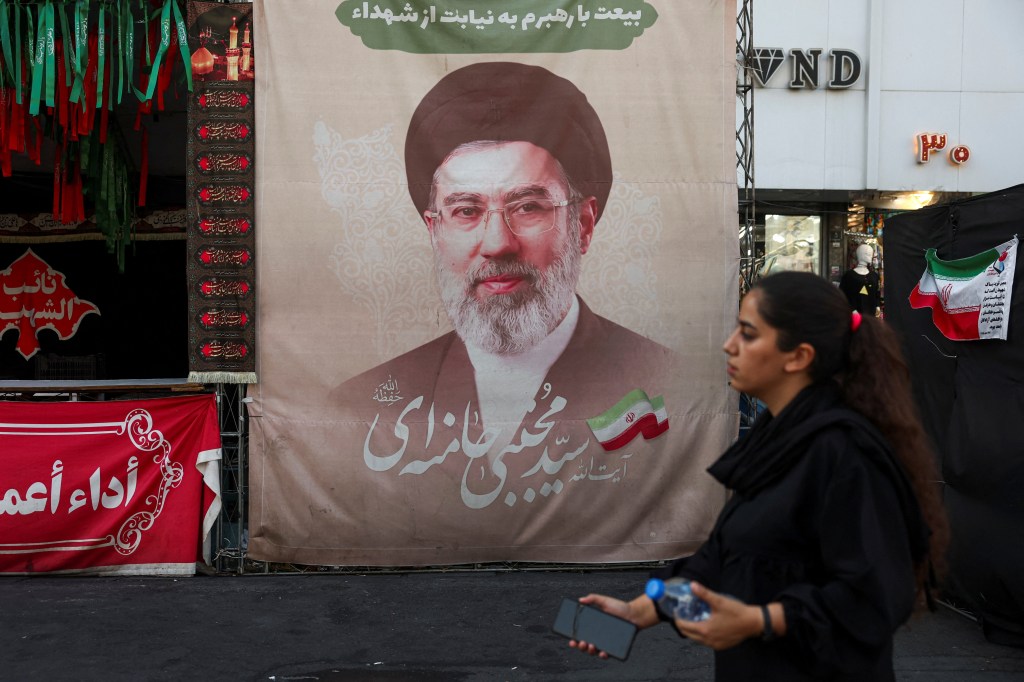 A woman in black walks past a large banner featuring Iran's Supreme Leader Mojtaba Khamenei in Tehran.