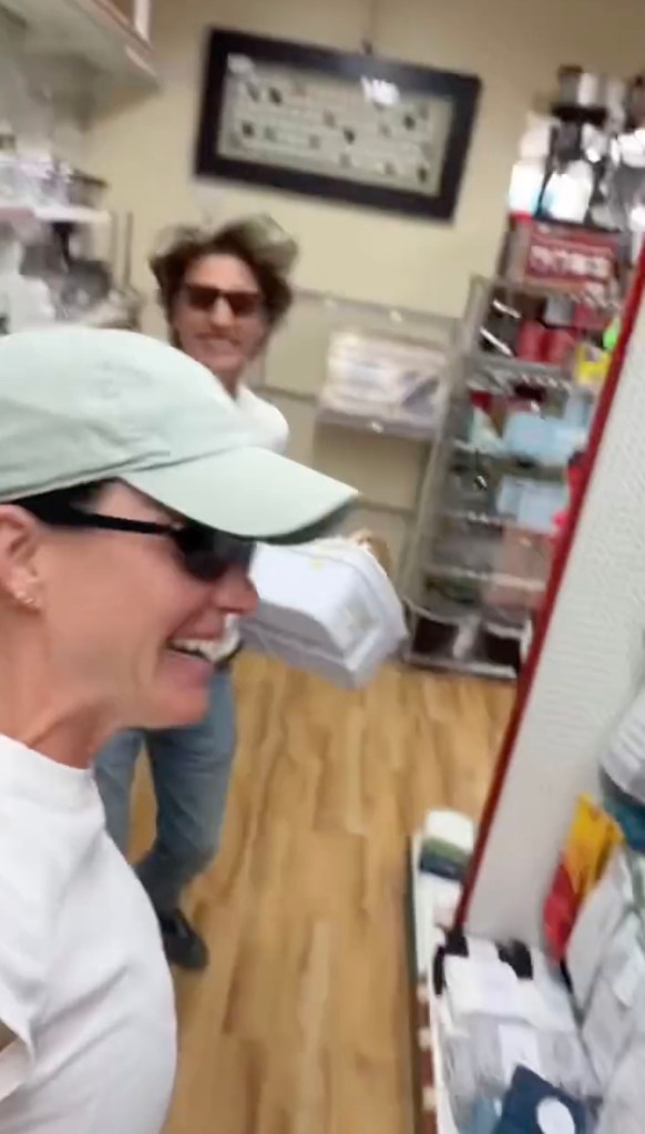 Katy Perry, wearing a cap and sunglasses, laughs as Justin Trudeau, holding a toilet plunger, smiles in a hardware store.