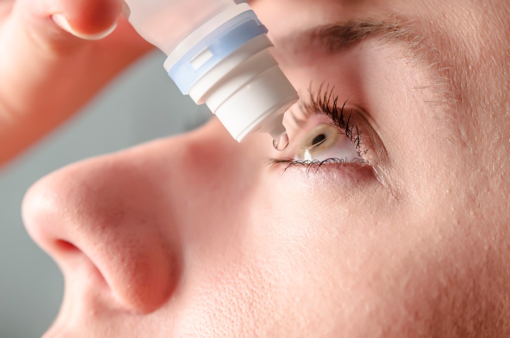 A close-up of a young woman applying an eye drop to her left eye.
