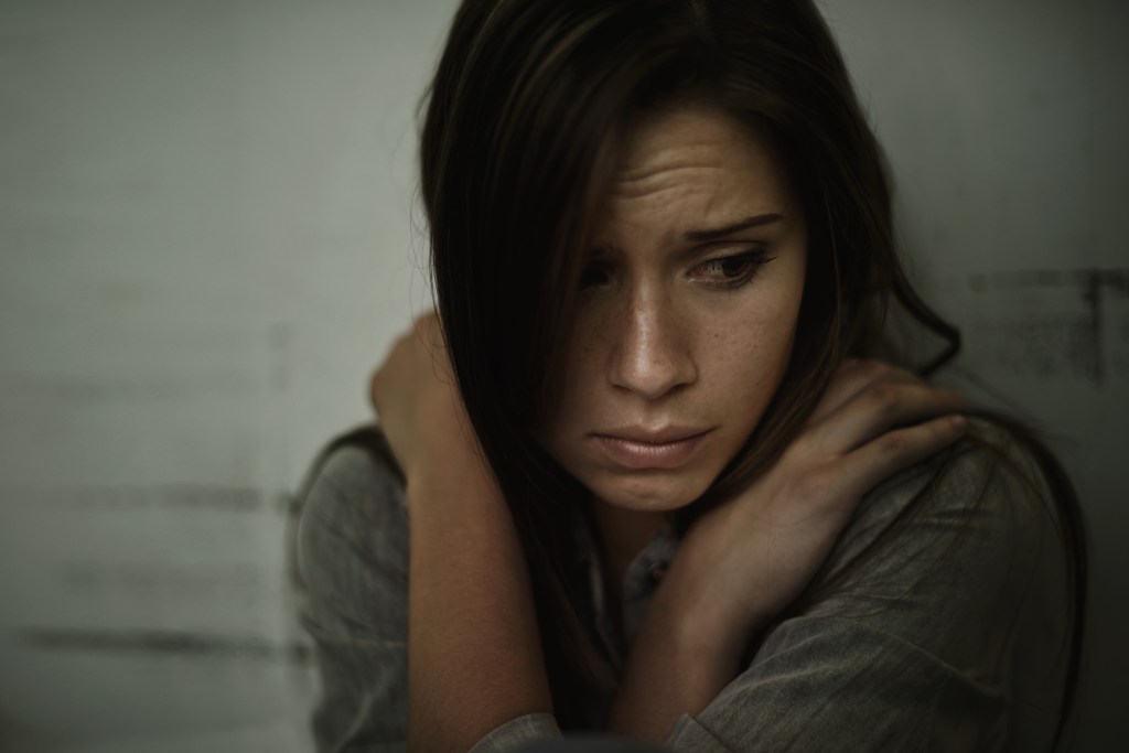 A young woman with a fearful and anxious expression, hugging herself, with dark hair cascading over one side of her face.