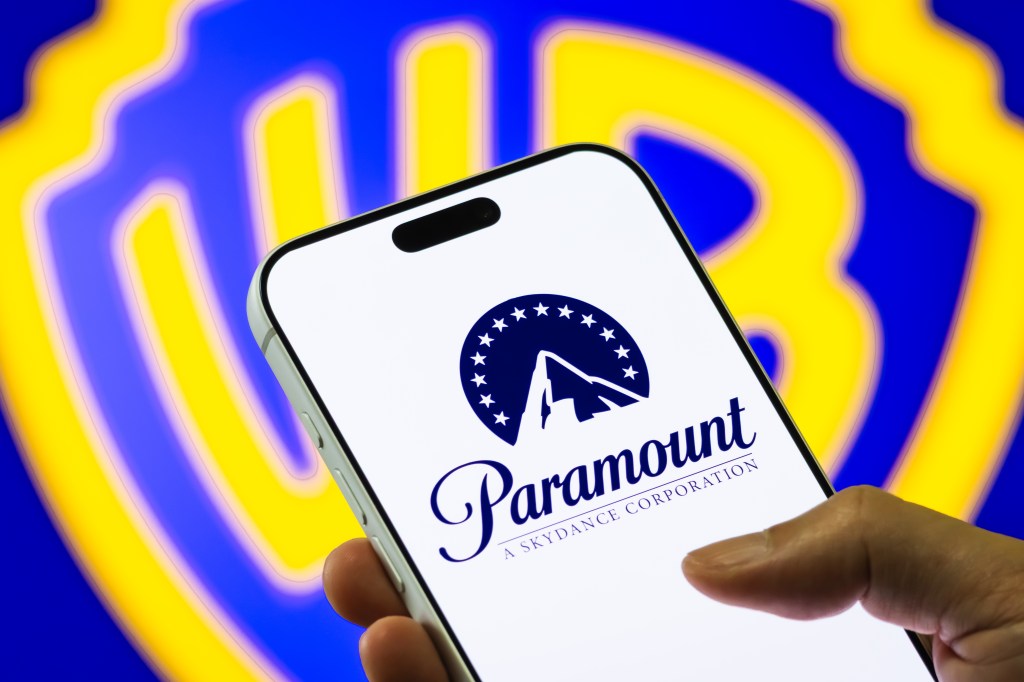 Photo illustration of a smartphone displaying the Paramount Skydance logo in front of a blurred Warner Bros. Discovery emblem.