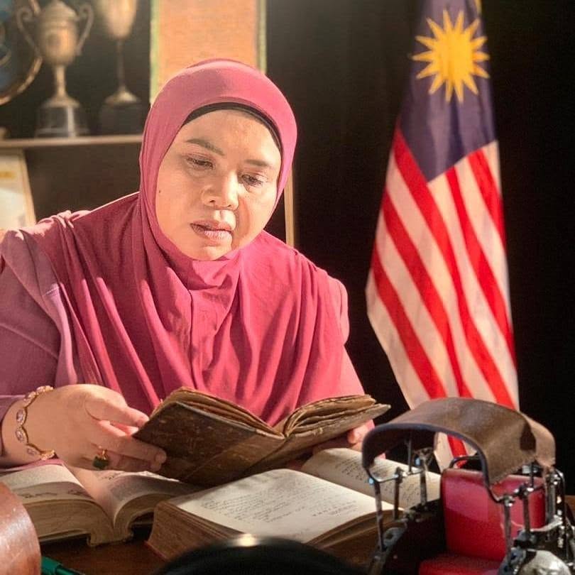 Professor Solehah Yaacob reading a book at a desk with a Malaysian flag behind her.