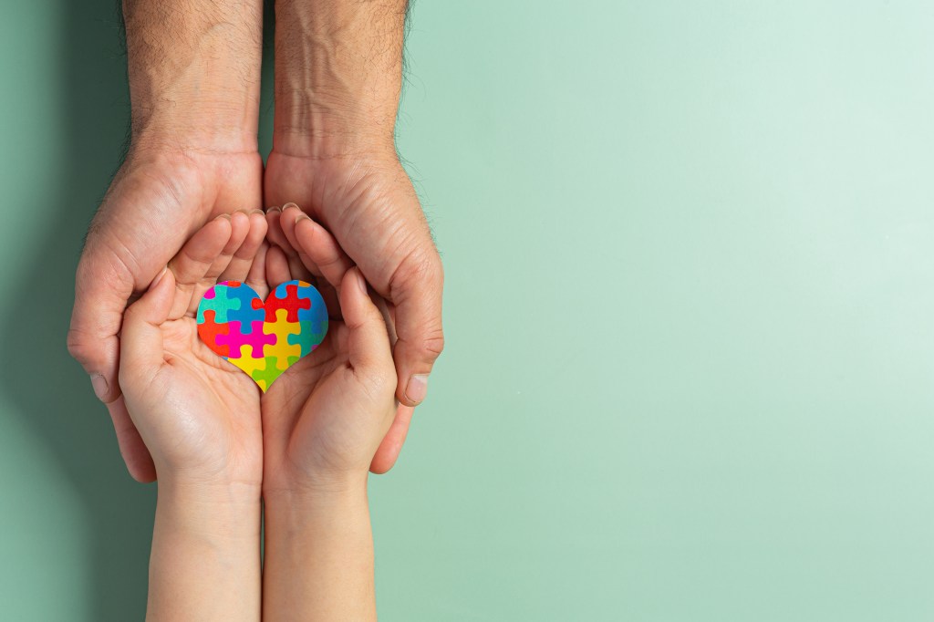 Adult hands holding a child's hands, which in turn hold a colorful heart-shaped jigsaw puzzle.