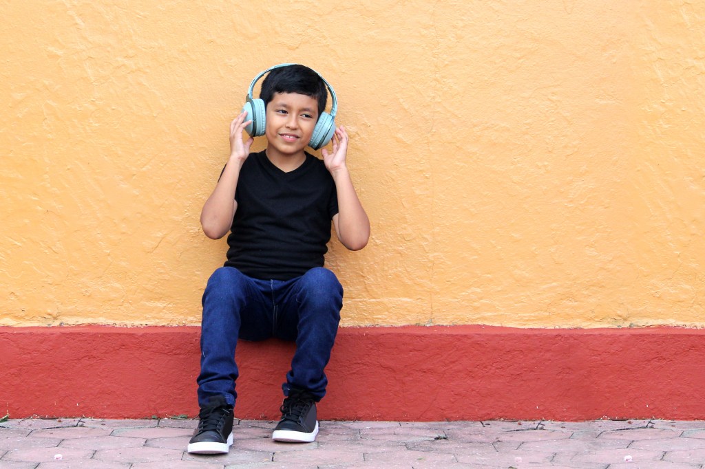 Brown boy wearing anti-noise headphones sitting against an orange and red wall.