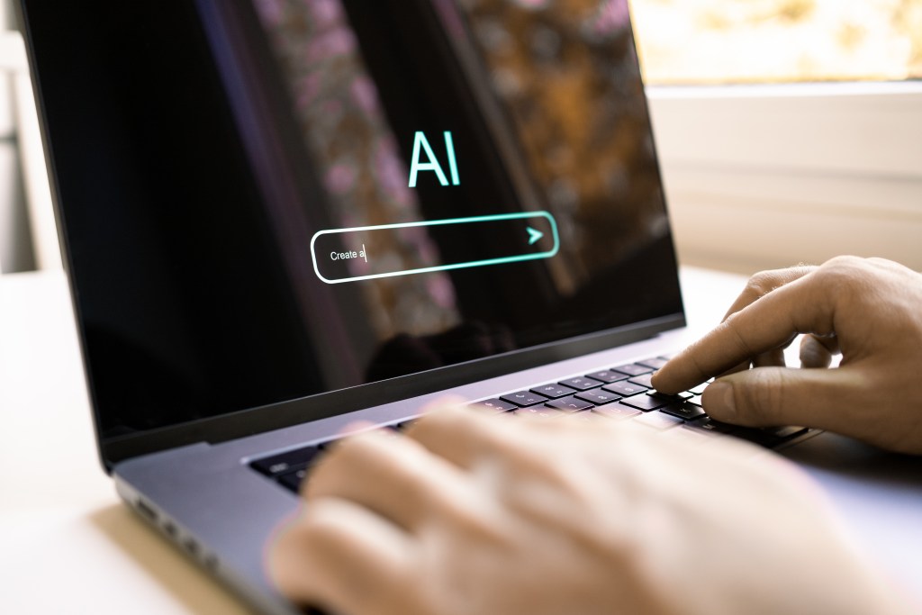 Hands typing on a laptop with "AI" and a "Create a" prompt visible on the screen.