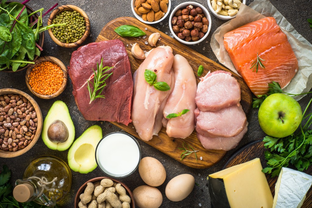 A top-down view of various protein sources including raw beef, chicken, pork, salmon, nuts, beans, lentils, cheese, eggs, milk, avocado, spinach, and oil on a dark table.