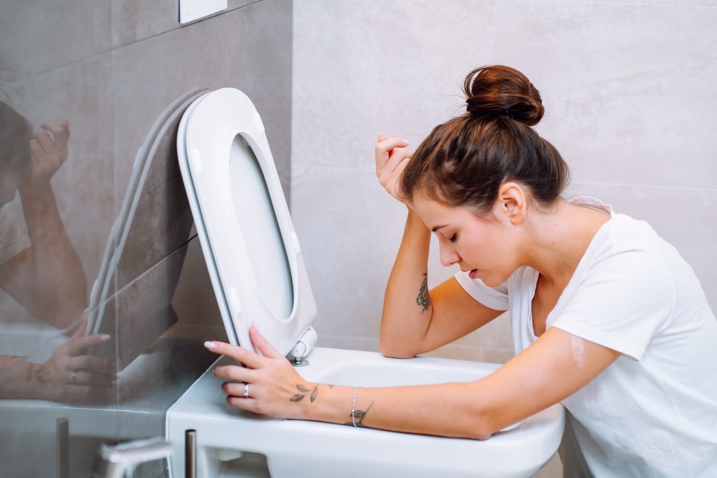 A young woman with her hair in a bun leans over a toilet in a bathroom.