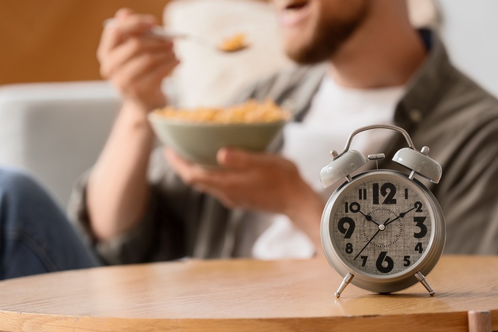 An alarm clock sits on a wooden table while a man in the background eats cereal.