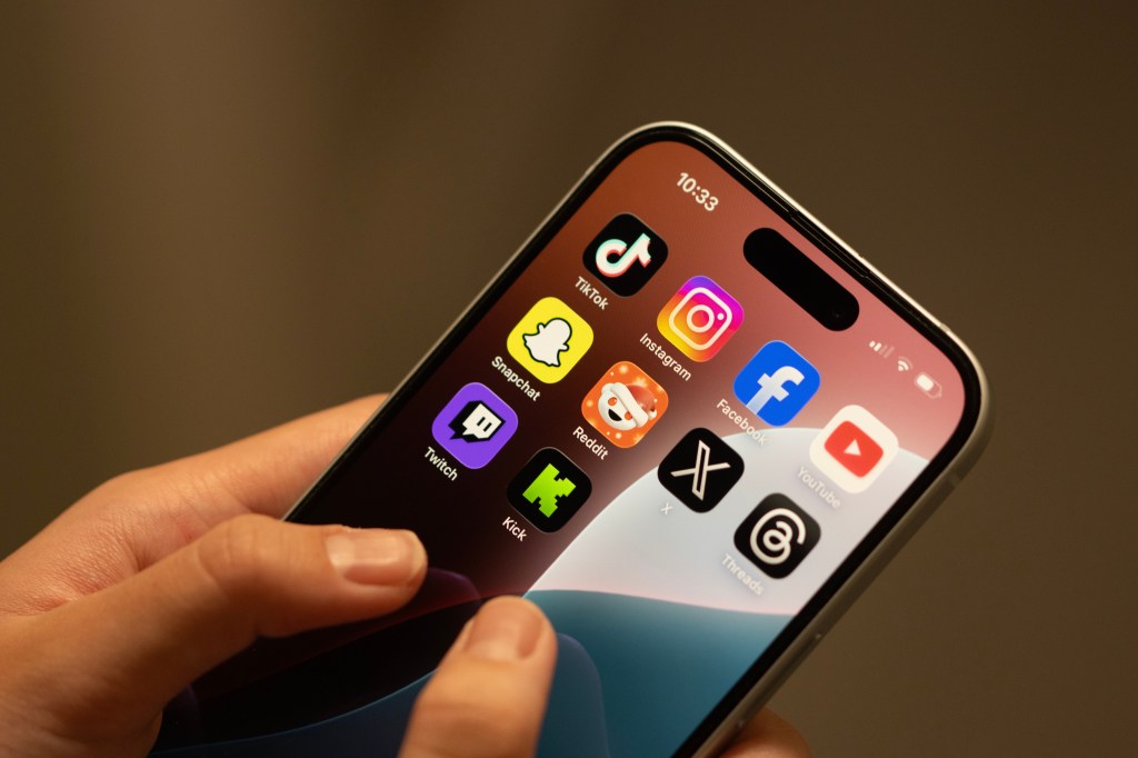 Hand holding an iPhone displaying app icons for TikTok, Instagram, Facebook, YouTube, Snapchat, Reddit, X, Threads, Twitch, and Kick.