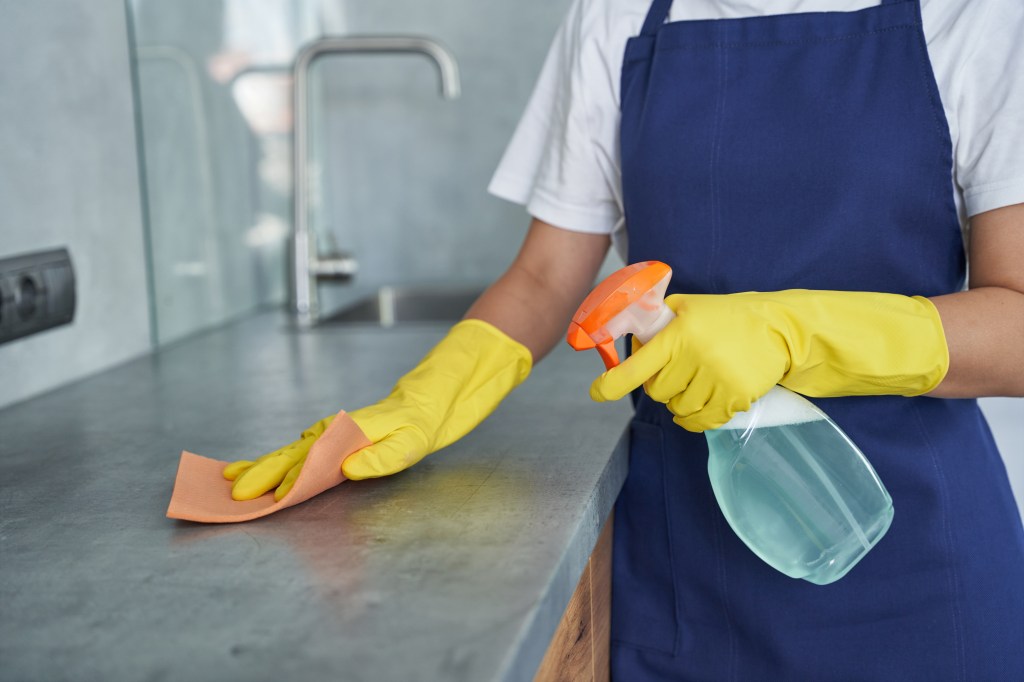 A person in yellow gloves and a blue apron spraying and wiping a kitchen countertop.