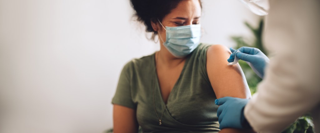 A masked woman receiving a COVID-19 vaccination in her arm from a healthcare worker wearing blue gloves.