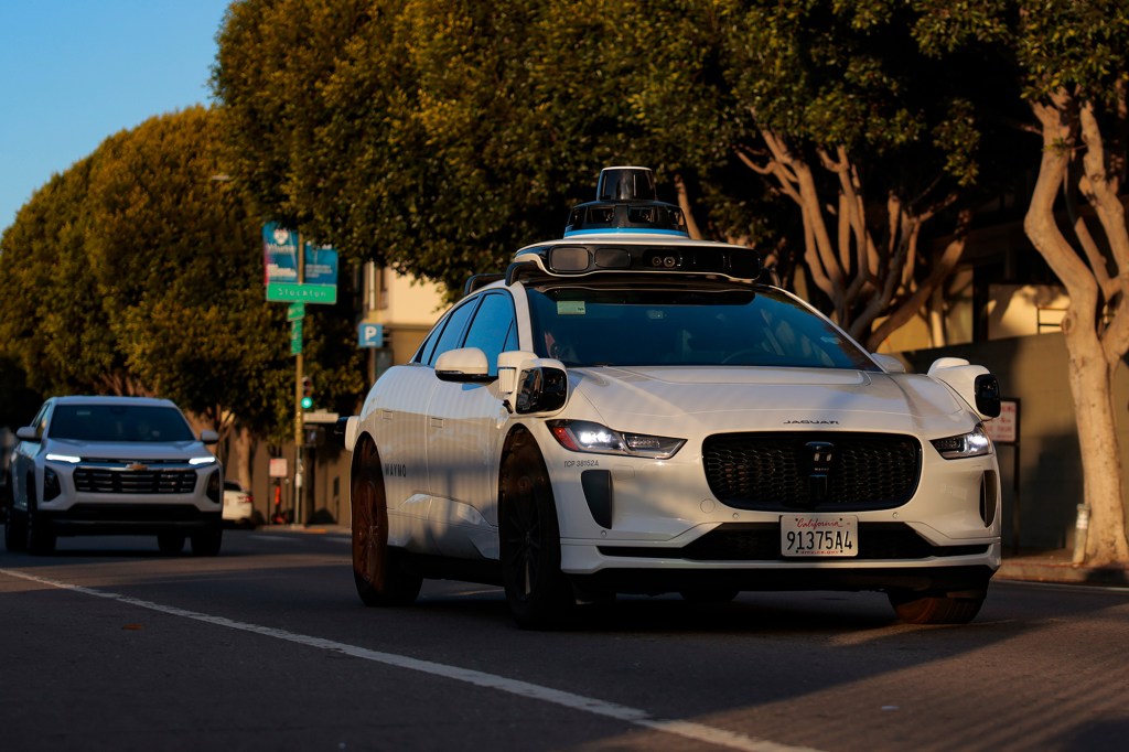 A white Jaguar Waymo autonomous car with roof-mounted sensors driving on a city street.