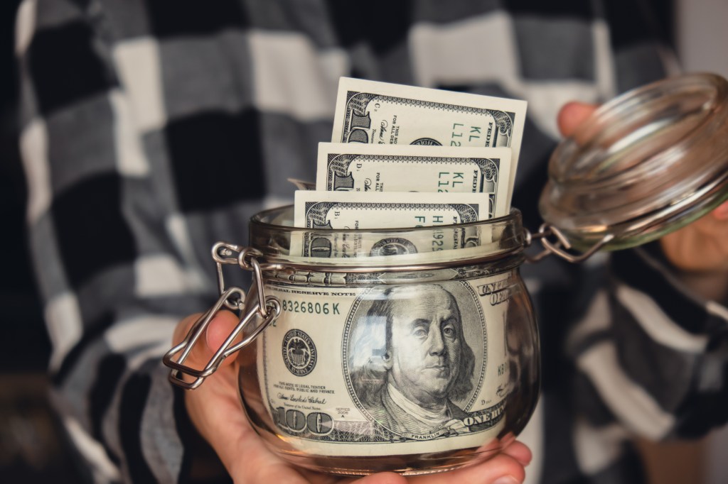 A person holding a glass jar filled with dollar banknotes.