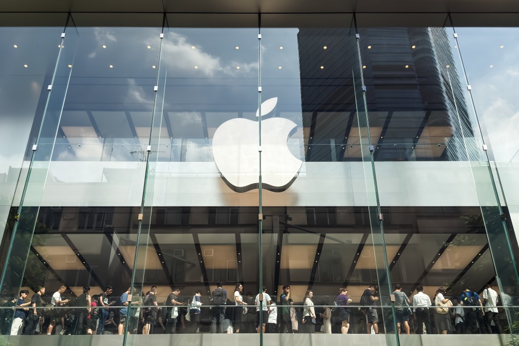 Customers queueing inside an Apple Store behind a large glass facade with the Apple logo.