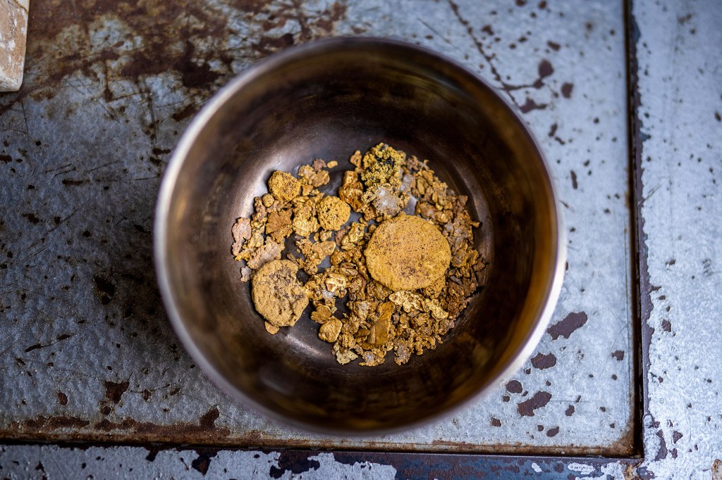 A metal bowl filled with raw gold nuggets and two flat, circular gold pieces, sitting on a rusted metal surface.