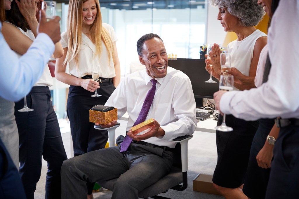 Coworkers celebrating a colleague's retirement in the office, with the retired colleague holding a gift box and smiling.
