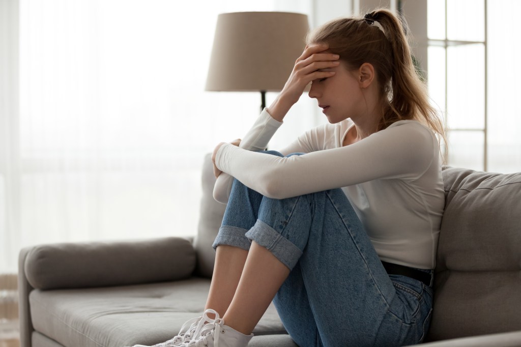 A sad woman sits on a couch, embracing her knees with her face covered in her hand.