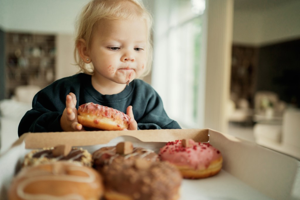 A toddler with frosting on her mouth and chin holds a pink iced donut over a box of other donuts.