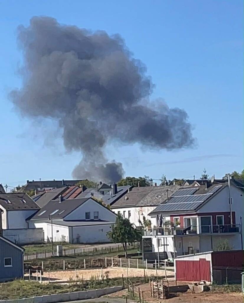 Thick black smoke rises above houses into a clear blue sky.