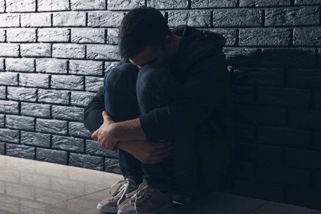 A depressed man sits huddled against a dark brick wall, contemplating suicide.