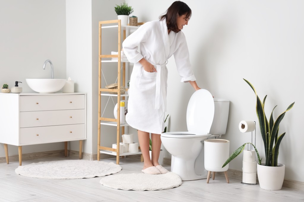 A young woman in a white bathrobe flushes a toilet in a modern bathroom.