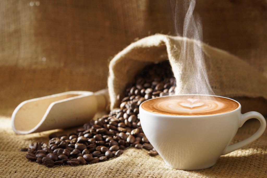 A white coffee cup with hot steaming latte art, surrounded by roasted coffee beans and a burlap sack.