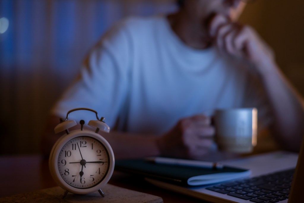 A person yawning while drinking coffee at a desk with an alarm clock and laptop.