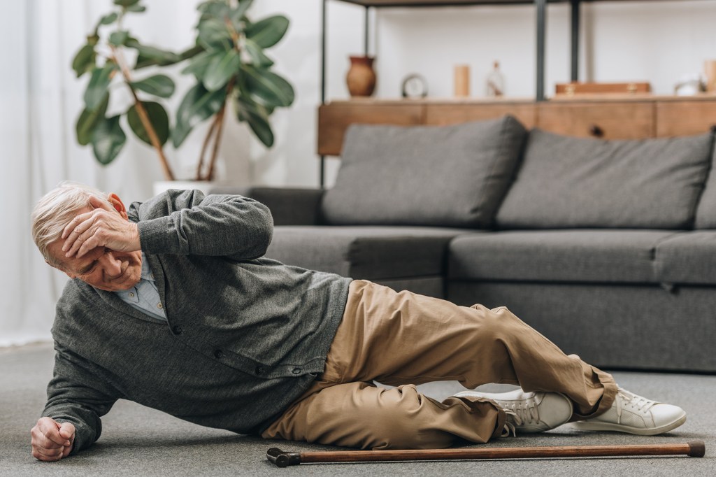 An elderly man lying on the floor holding his head, with a cane nearby.