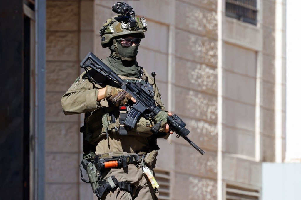An Israeli soldier in tactical gear, sunglasses, and a balaclava, carrying a rifle during a raid in Beddu.