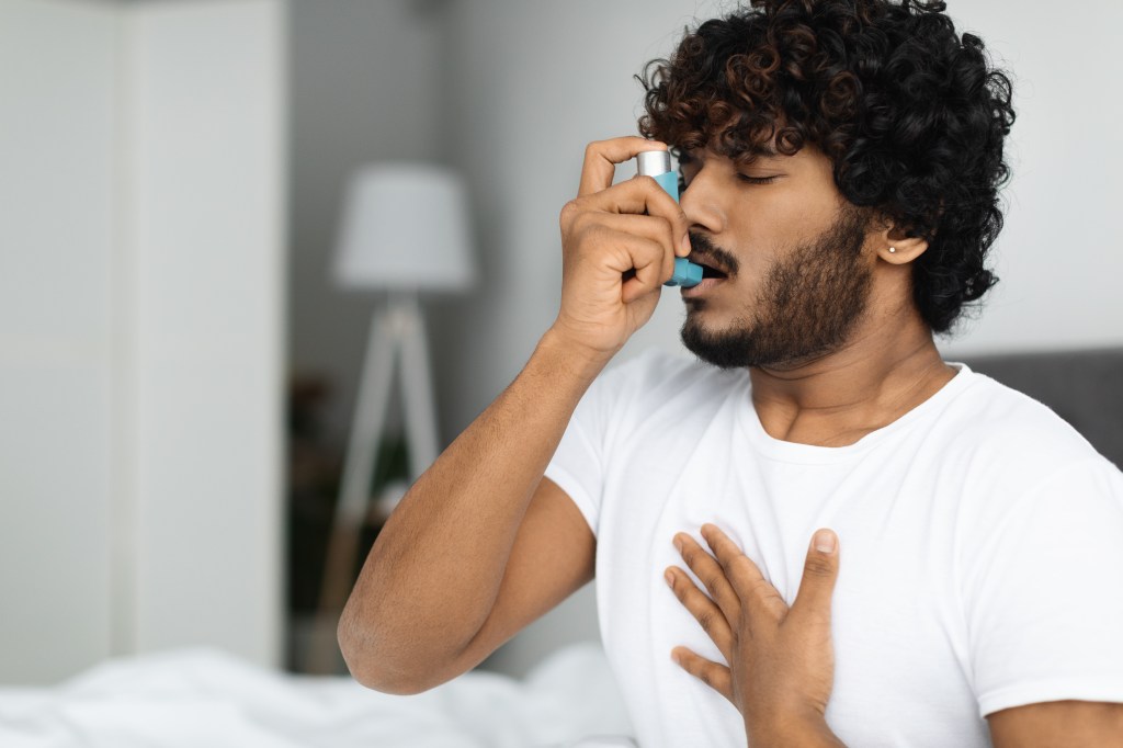 A man in a white shirt, with curly dark hair and a beard, uses an inhaler with his eyes closed and his other hand on his chest.