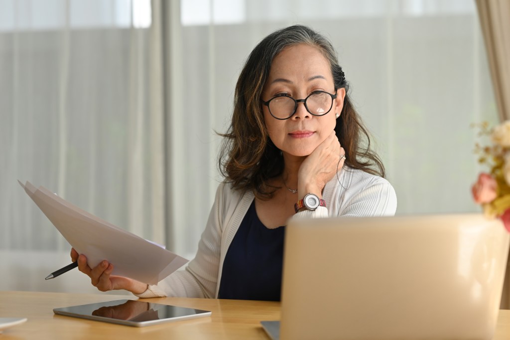 An elderly woman wearing glasses works on a laptop while sitting at a desk in a living room.