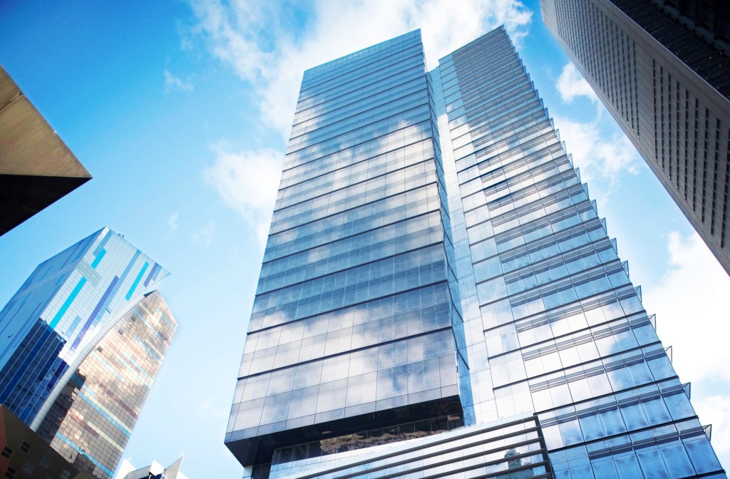 Looking up at two tall, glass-faced skyscrapers reflecting the blue sky and clouds.